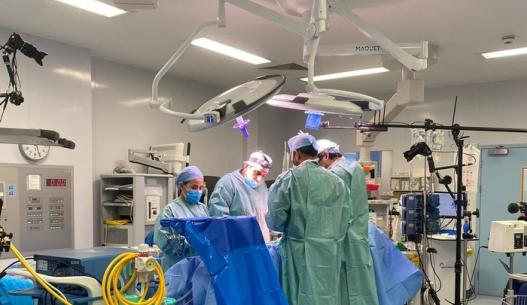 Interior of surgical theatre, surgeons in gowns and scrubs operate under bright lights. A documentary crew-member films from the far right of the image, sat on a chair
