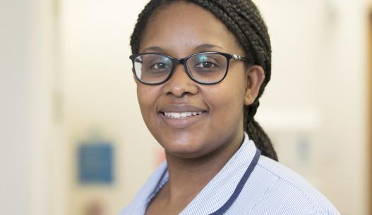 Staff Nurse Joyce Twene-Dove, a Black woman with glasses and box braids, smiles for the camera while wearing a pale blue uniform