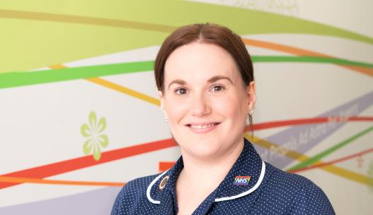 Heather Jones, Junior Sister at the OCCYP. Heather is a white woman with long brown hair. She is smiling in front of a colourful mural and is wearing a dark blue nurse's uniform.