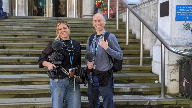 two members of the documentary crew pose on the hospital steps, exterior, holding video cameras and boom microphones