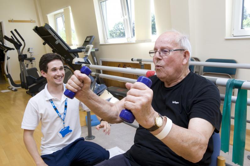 An older man lifts two small weights with the support of a physiotherapist