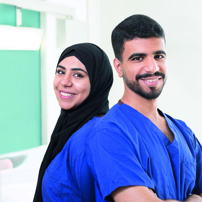 Royal Marsden Nurses Sara and Abed, brother and sister, stand back-to-back. Sara is wearing a black hijab and Abed is dressed in blue scrubs.